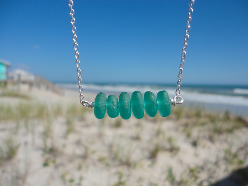 May include: A silver chain necklace with a row of six teal sea glass beads. The necklace is photographed against a blurred background of a beach.