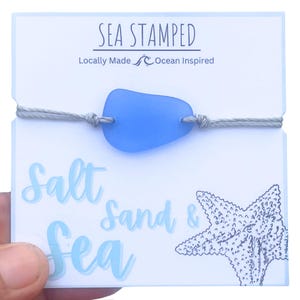 Peut inclure: Un bracelet en verre de mer bleu clair avec un cordon gris et une carte blanche qui dit "Sea Stamped" et "Locally Made Ocean Inspired" et "Salt Sand & Sea" avec une illustration d'&eacute;toile de mer noire et blanche.