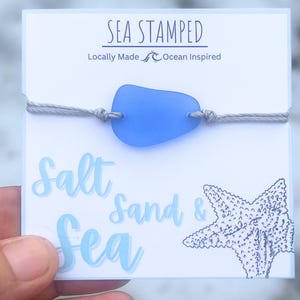 May include: A light blue sea glass charm bracelet with a gray cord and a white card that says "Sea Stamped" "Locally Made Ocean Inspired" and "Salt Sand & Sea".