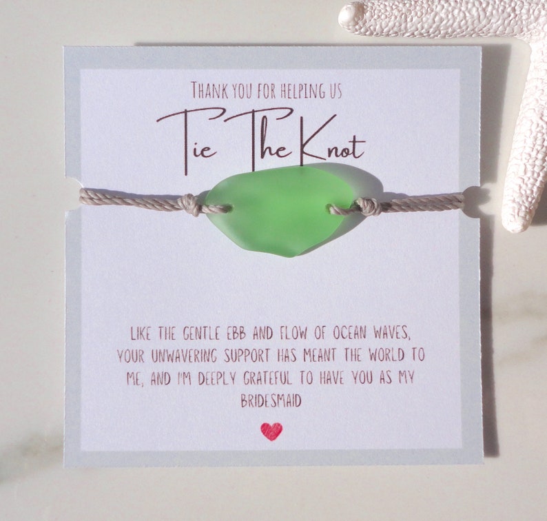 Puede incluir: Una tarjeta blanca con el texto "Gracias por ayudarnos a atar el nudo" y un amuleto de vidrio de mar verde en una cuerda beige. La tarjeta tambi&eacute;n incluye el texto "Como el suave vaiv&eacute;n de las olas del oc&eacute;ano, tu apoyo inquebrantable ha significado el mundo para m&iacute;, y estoy profundamente agradecida de tenerte como mi dama de honor."