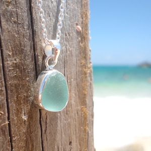 May include: A silver chain necklace with a light blue sea glass pendant. The pendant is set in a silver bezel and hangs against a wooden background.
