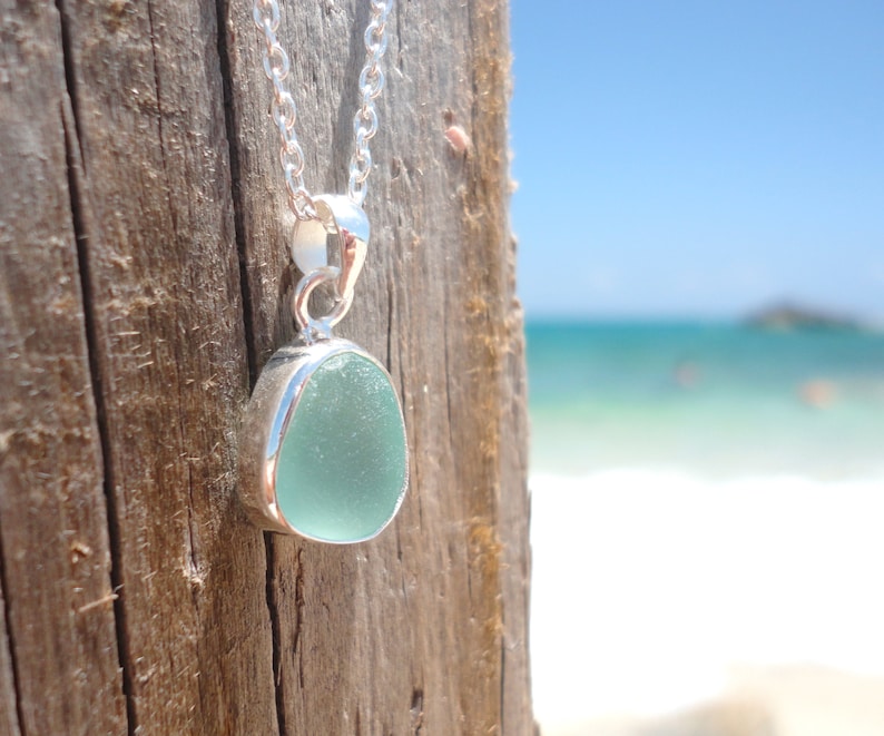 May include: A silver chain necklace with a light blue sea glass pendant. The pendant is set in a silver bezel and hangs against a wooden background.