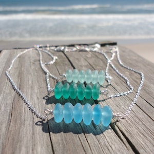 May include: Three silver necklaces with a row of light blue and green sea glass beads. The necklaces are displayed on a wooden surface.