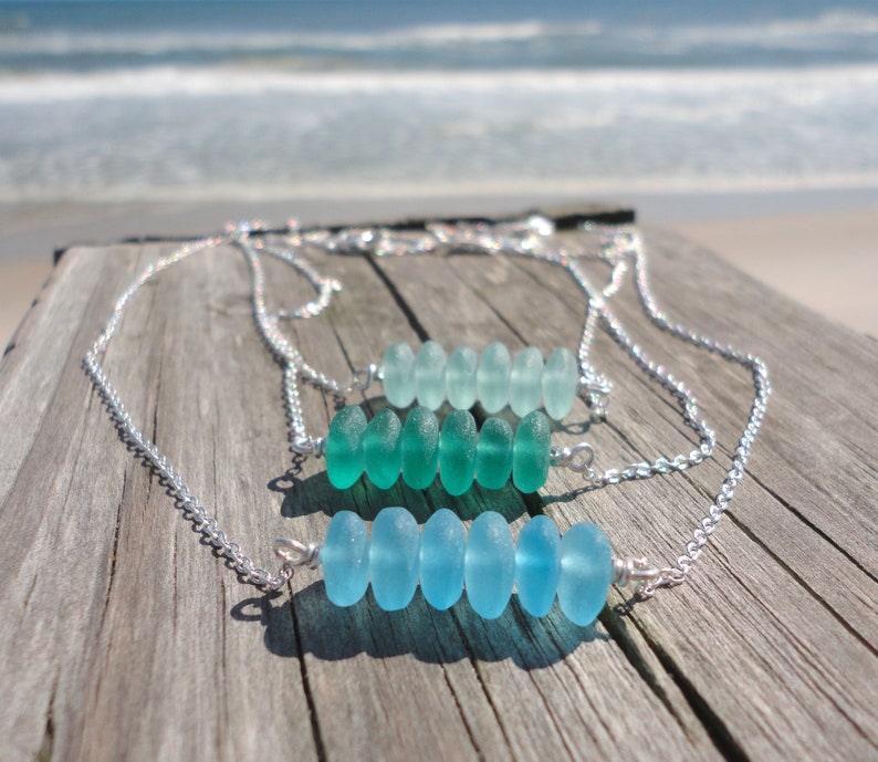 May include: Three silver necklaces with a row of light blue and green sea glass beads. The necklaces are displayed on a wooden surface.