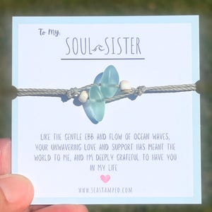 Może przedstawiać: Bransoletka ze sznurka w kolorze szarym z niebieskimi i białymi koralikami. Bransoletka znajduje się na kartce z tekstem "To my SOUL SISTER" i wiadomością o przypływie i odpływie oceanicznych fal. Na kartce znajduje się r&oacute;wnież adres strony internetowej "www.seastamped.com".