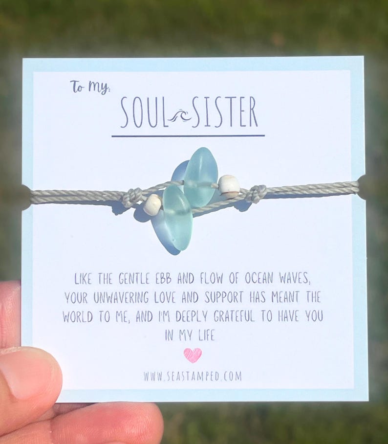 Może przedstawiać: Bransoletka ze sznurka w kolorze szarym z niebieskimi i białymi koralikami. Bransoletka znajduje się na kartce z tekstem "To my SOUL SISTER" i wiadomością o przypływie i odpływie oceanicznych fal. Na kartce znajduje się r&oacute;wnież adres strony internetowej "www.seastamped.com".