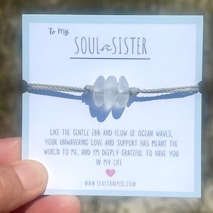 Może przedstawiać: Bransoletka ze sznurka w kolorze szarym z trzema białymi koralikami ze szkła morskiego. Bransoletka jest prezentowana na kartce z tekstem "To my SOUL SISTER" i wiadomością o przypływie i odpływie fal oceanu. Na kartce znajduje się r&oacute;wnież adres strony internetowej "www.seastamped.com".