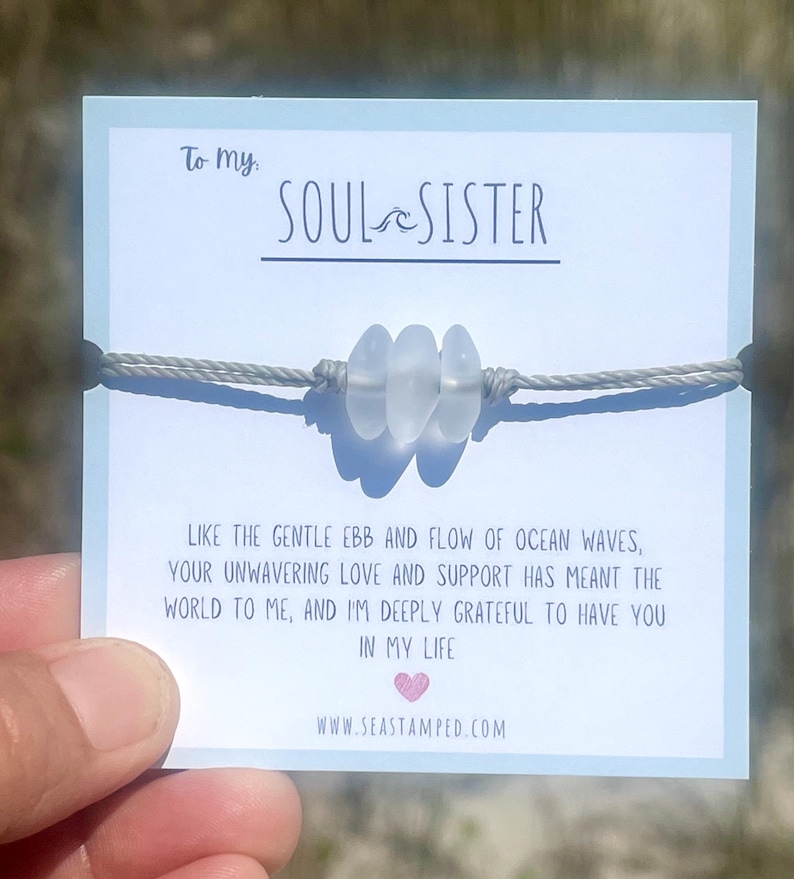 Może przedstawiać: Bransoletka ze sznurka w kolorze szarym z trzema białymi koralikami ze szkła morskiego. Bransoletka jest prezentowana na kartce z tekstem "To my SOUL SISTER" i wiadomością o przypływie i odpływie fal oceanu. Na kartce znajduje się r&oacute;wnież adres strony internetowej "www.seastamped.com".