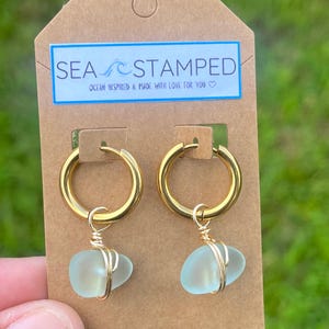 May include: Gold hoop earrings with sea glass pendants. The earrings are attached to a brown tag with the text "SEA STAMPED" and "OCEAN INSPIRED & MADE WITH LOVE FOR YOU". The sea glass is a light blue color.