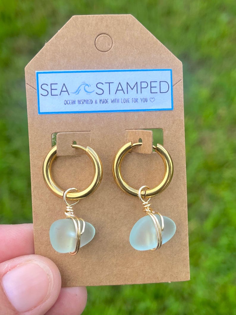 May include: Gold hoop earrings with sea glass pendants. The earrings are attached to a brown tag with the text "SEA STAMPED" and "OCEAN INSPIRED & MADE WITH LOVE FOR YOU". The sea glass is a light blue color.