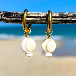 May include: Gold hoop earrings with white and tan seashell-like beads. The earrings have a small pink bead and a gold-colored accent at the bottom of the white bead. The background is a blurred view of the ocean and sky.