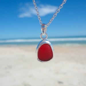 May include: A silver necklace with a red sea glass pendant. The pendant is shaped like a teardrop and has a smooth, polished surface.