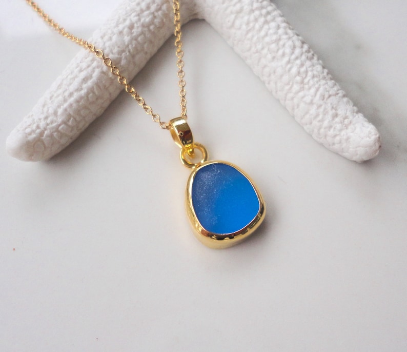 May include: A gold chain necklace with a blue sea glass pendant. The pendant is a teardrop shape with a smooth, polished surface.