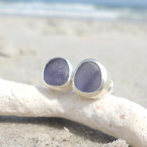 May include: A pair of stud earrings featuring smooth, lavender-colored sea glass set in silver bezels. The earrings are displayed on a piece of driftwood against a sandy beach background, with the ocean visible in the distance.