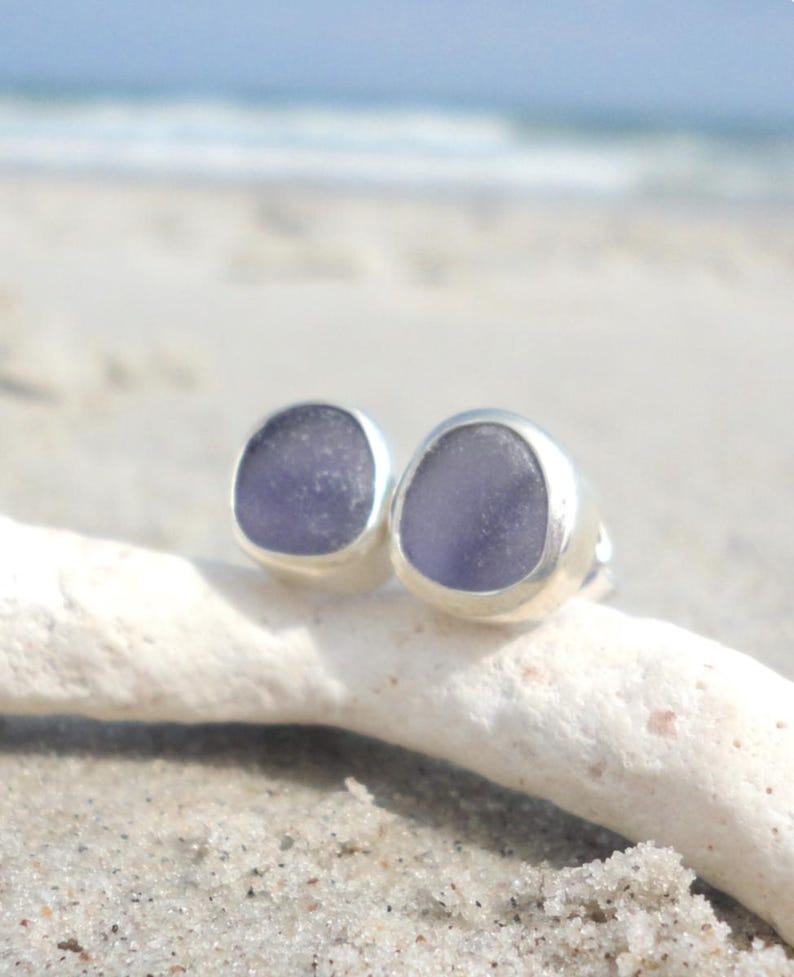 May include: A pair of stud earrings featuring smooth, lavender-colored sea glass set in silver bezels. The earrings are displayed on a piece of driftwood against a sandy beach background, with the ocean visible in the distance.