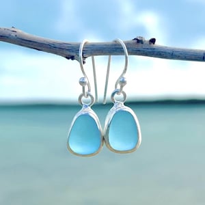 May include: A pair of light blue teardrop-shaped earrings with a silver border, hanging from silver hooks. The earrings are suspended from a small branch against a blurred background of the ocean and sky.