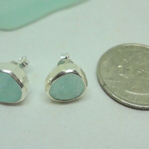 May include: A pair of sea glass stud earrings with a quarter for size comparison. The earrings feature a light blue, rounded sea glass stone set in a silver bezel. The earrings are small and delicate, perfect for everyday wear.