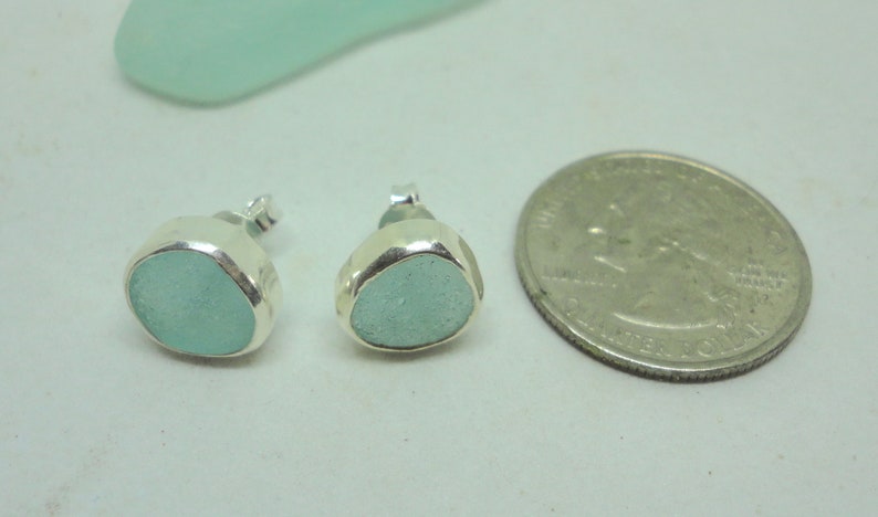 May include: A pair of sea glass stud earrings with a quarter for size comparison. The earrings feature a light blue, rounded sea glass stone set in a silver bezel. The earrings are small and delicate, perfect for everyday wear.