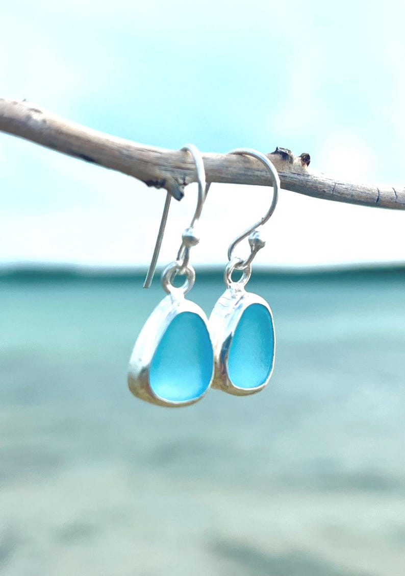 May include: A pair of silver-toned drop earrings with teardrop-shaped, light blue sea glass stones. The earrings are suspended from a thin, curved branch against a blurred blue background.