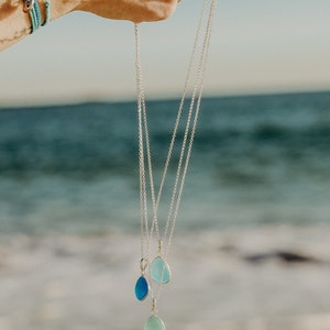 Op de afbeelding: Drie zilveren kettingen met traanvormige hangers van zeeglas. De hangers zijn in verschillende tinten blauw en groen.