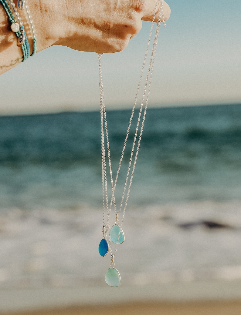 Op de afbeelding: Drie zilveren kettingen met traanvormige hangers van zeeglas. De hangers zijn in verschillende tinten blauw en groen.
