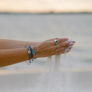 Peut inclure: Une main portant plusieurs bracelets, tenant du sable et le laissant tomber entre ses doigts. La personne porte une bague avec une pierre bleue.
