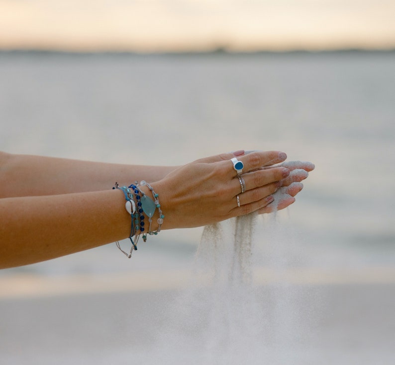 Peut inclure: Une main portant plusieurs bracelets, tenant du sable et le laissant tomber entre ses doigts. La personne porte une bague avec une pierre bleue.