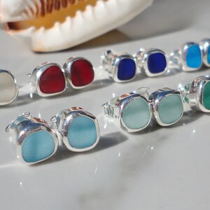 May include: A collection of colorful stud earrings with square-shaped sea glass set in silver. The earrings come in various colors, including white, red, blue, turquoise, green, and purple. A seashell and starfish are in the background.