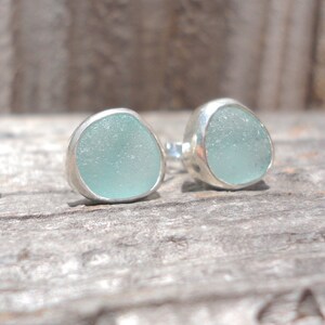 May include: A pair of stud earrings featuring pale blue sea glass set in silver bezels. The sea glass has a smooth, rounded shape. The earrings are displayed on a weathered wooden surface.