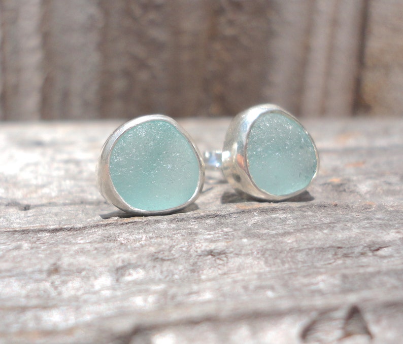 May include: A pair of stud earrings featuring pale blue sea glass set in silver bezels. The sea glass has a smooth, rounded shape. The earrings are displayed on a weathered wooden surface.