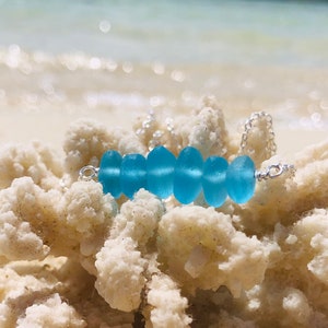 May include: A silver chain necklace with a row of light blue sea glass beads. The necklace is resting on a piece of white coral.