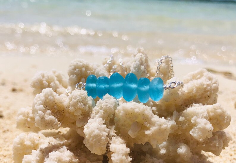 May include: A silver chain necklace with a row of light blue sea glass beads. The necklace is resting on a piece of white coral.