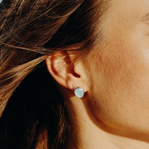 May include: A close-up of a light blue square stud earring. The earring is set against the backdrop of a person's ear and hair, with the focus on the earring's shape and color. The earring is a simple, minimalist design.