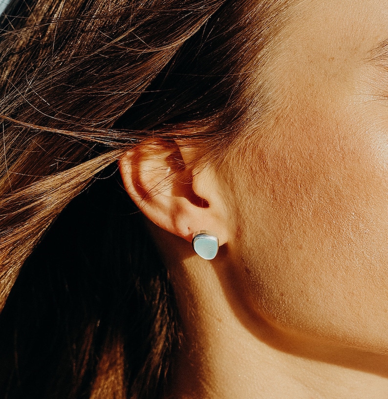May include: A close-up of a light blue square stud earring. The earring is set against the backdrop of a person's ear and hair, with the focus on the earring's shape and color. The earring is a simple, minimalist design.