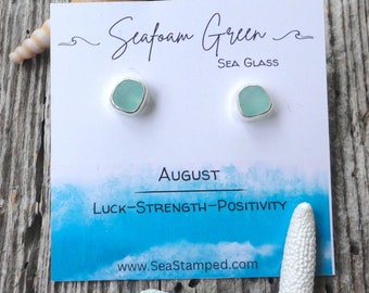 Może przedstawiać: Kolczyki sztyfty z zielonego szkła morskiego z srebrnymi oprawami. Kolczyki są prezentowane na karcie z napisem "Seafoam Green Sea Glass" i "August Luck-Strength-Positivity". Na zdjęciu znajduje się również muszla i rozgwiazda.