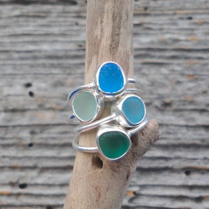 May include: Four silver rings with different coloured sea glass stones. The rings are stacked on a piece of driftwood.