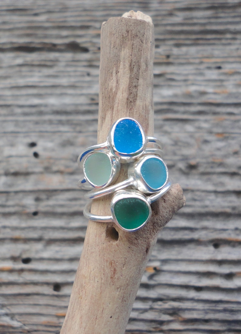 May include: Four silver rings with different coloured sea glass stones. The rings are stacked on a piece of driftwood.