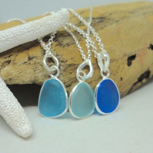 Op de afbeelding: Drie zilveren kettingen met hangers van zeeglas. De hangers zijn in tinten blauw, van lichtblauw tot donkerblauw.