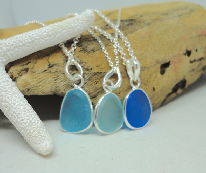 Op de afbeelding: Drie zilveren kettingen met hangers van zeeglas. De hangers zijn in tinten blauw, van lichtblauw tot donkerblauw.