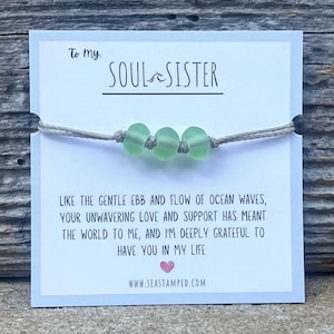 Może przedstawiać: Bransoletka ze sznurka w jasnoszarym kolorze z trzema jasnozielonymi szklanymi koralikami. Bransoletka jest przymocowana do kartki z tekstem "To my SOUL SISTER" i wiadomością o znaczeniu przyjaźni. Na kartce znajduje się r&oacute;wnież adres strony internetowej "www.seastamped.com".