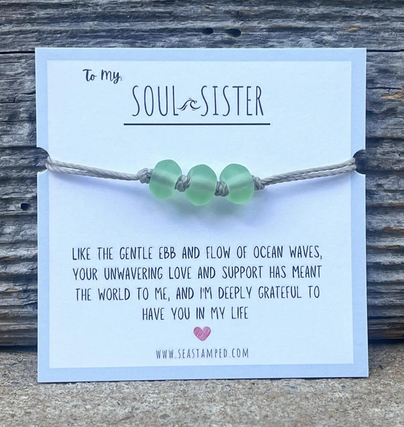 Może przedstawiać: Bransoletka ze sznurka w jasnoszarym kolorze z trzema jasnozielonymi szklanymi koralikami. Bransoletka jest przymocowana do kartki z tekstem "To my SOUL SISTER" i wiadomością o znaczeniu przyjaźni. Na kartce znajduje się r&oacute;wnież adres strony internetowej "www.seastamped.com".