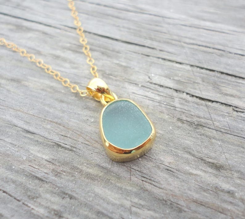 May include: A gold chain necklace with a light blue sea glass pendant. The pendant is set in a gold bezel.