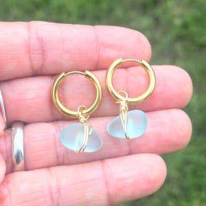 May include: A pair of gold-colored hoop earrings, each featuring a light blue, sea glass pendant. The sea glass is wire-wrapped in gold-colored wire and hangs from the bottom of the hoop. The background is a blurred green.