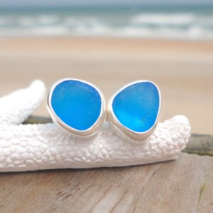May include: A pair of blue sea glass stud earrings with a silver bezel setting. The earrings are displayed on a white, textured surface, with a blurred beach and ocean background.