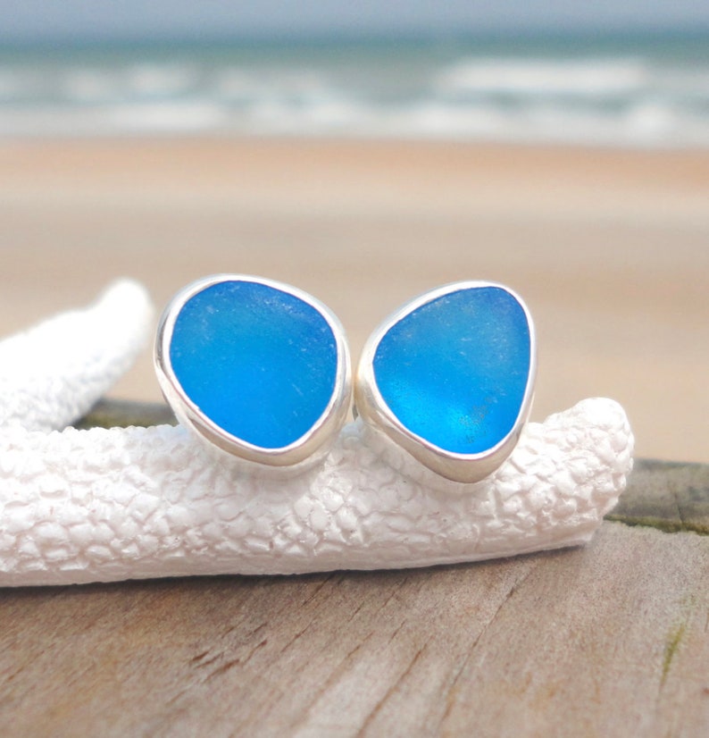 May include: A pair of blue sea glass stud earrings with a silver bezel setting. The earrings are displayed on a white, textured surface, with a blurred beach and ocean background.