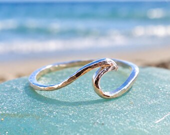 May include: A silver ring featuring a continuous, hammered metal band shaped into a stylised ocean wave. The surface of the metal has a textured, reflective finish that catches the light, creating a distinct wave silhouette across the finger.