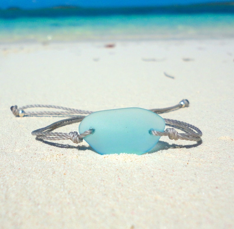 Peut inclure: Un bracelet en corde grise avec un charme en verre de mer bleu clair. Le bracelet est pos&eacute; sur une plage de sable blanc.