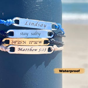 Puede incluir: Cuatro pulseras personalizadas con etiquetas metálicas rectangulares y cordones anudados. Las etiquetas están grabadas con los nombres "Lindsay", "stay salty", coordenadas y "Matthew 5:13". Las pulseras son de varios colores, incluyendo azul, negro, dorado y beige. La palabra "Waterproof" también es visible.