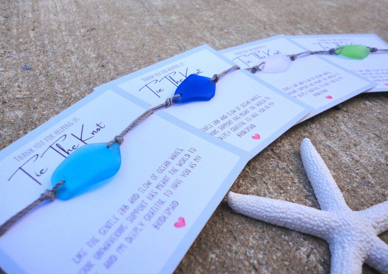 Puede incluir: Un conjunto de cuatro pulseras de recuerdo con abalorios de vidrio de mar y un mensaje de agradecimiento por ayudar a atar el nudo. El mensaje dice: "Gracias por ayudarnos a atar el nudo. Como el suave vaiv&eacute;n de las olas del oc&eacute;ano, tu apoyo inquebrantable ha significado el mundo para nosotros. Te estoy profundamente agradecido por tenerte como mi dama de honor."