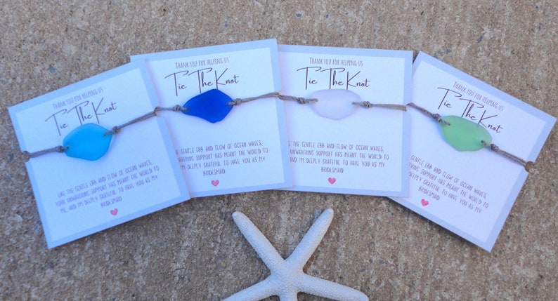 Puede incluir: Cuatro cajas de regalo con pulseras de vidrio de mar y tarjetas que dicen "Gracias por ayudarnos a atar el nudo". Las tarjetas tienen un mensaje sobre el suave vaiv&eacute;n de las olas del oc&eacute;ano y el apoyo inquebrantable que ha significado el mundo para la pareja. Las cajas est&aacute;n sobre una superficie arenosa con una estrella de mar blanca.